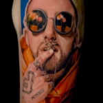 Фото тату на руке с изображением Mac Miller в оранжевом капюшоне и с сигаретой в руках. Рисунок татуировки с использованием ярких цветов и детальной проработкой.