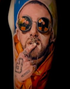 Фото тату на руке с изображением Mac Miller в оранжевом капюшоне и с сигаретой в руках. Рисунок татуировки с использованием ярких цветов и детальной проработкой.