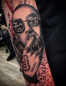 Фото тату на руке с портретом Mac Miller в темных очках и с загадочным выражением лица. Рисунок татуировки с использованием черно-белой гаммы и красных акцентов.