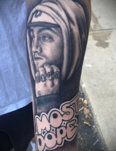 Фото тату на руке с изображением Mac Miller в капюшоне и с кольцами на пальцах. Черно-белый рисунок татуировки.