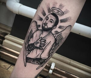 Фото тату на руке с изображением Mac Miller в образе ангела с крыльями и нимбом. Черно-белый рисунок татуировки.