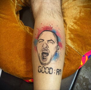 Фото тату: портрет Mac Miller с открытым ртом в крике, с брызгами краски и надписью 'GOOD:AM'. Рисунок татуировки на руке в ярких цветах.