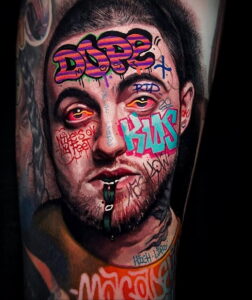 Фото тату: портрет Mac Miller, покрытый яркими надписями и рисунками в стиле граффити. Детализированный рисунок татуировки на руке.