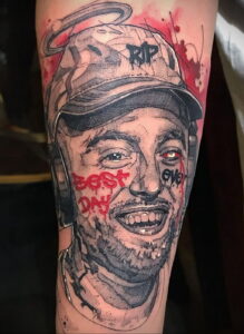 Фото тату: портрет Mac Miller в кепке с красными брызгами краски и надписью 'RIP'. Эмоциональный рисунок татуировки на руке, выражающий скорбь.