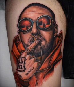Фото тату: реалистичный портрет Mac Miller в оранжевой куртке и круглых красных очках, держащего сигарету. Детализированный рисунок татуировки на предплечье.