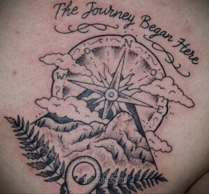 Фото тату на груди: компас, горы и облака с надписью "The Journey Began Here", выполненное в стиле олдскул.