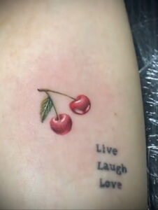 Фото тату: маленькая татуировка с вишней и надписью "Live Laugh Love" на запястье. Нежный эскиз татуировки для девушек.