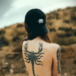 Фото тату скорпиона на спине девушки. Черный рисунок, детализированные клешни и хвост. Татуировка выглядит стильно и загадочно.