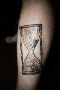 Фото тату песочных часов в черно-белых тонах с изображением силуэта женщины и черепа. Минималистичный дизайн, выразительный сюжет.