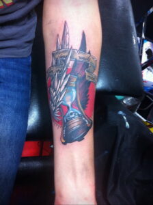 Фотография цветной татуировки на предплечье с изображением песочных часов, украшенных крыльями и лентами с надписями 'Time for me' и 'Time waits for man'. Яркий и символичный рисунок.