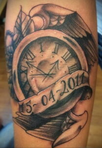 Фото татуировки на руке: карманные часы с датой '15-04-2011' и крыльями, выполненное в черно-серых тонах.