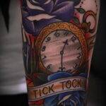 Фото тату на руке с изображением синих роз, карманных часов и надписи "Tick tock". Красивый и детализированный рисунок татуировки.
