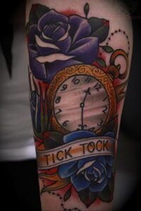 Фото тату на руке с изображением синих роз, карманных часов и надписи "Tick tock". Красивый и детализированный рисунок татуировки.