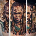 Фотография реалистичной татуировки на плече с изображением африканского воина с дредами и традиционными украшениями. Детальная проработка кожи и волос.