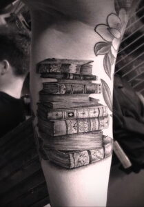 Фото тату на руке: реалистичная стопка старинных книг, перевязанных лентой, с добавлением цветочных элементов.