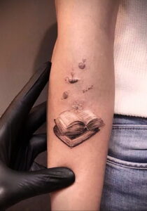 Фото тату на руке с изображением открытой книги, яблока, свечи и формулы E=mc². Детализированный рисунок татуировки в реалистичном стиле.