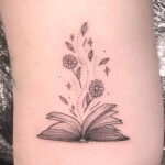 Фото тату на руке с изображением открытой книги, из которой растут цветы и искры. Минималистичный и элегантный рисунок татуировки.