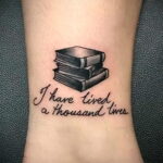 Фото тату на запястье с изображением небольшой стопки книг и надписью 'I have lived a thousand lives'. Простой и элегантный рисунок татуировки.