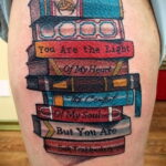 Фото тату на бедре с изображением стопки книг разных цветов и надписью 'You Are the Light Of My Heart'. Детализированный рисунок татуировки.