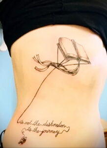 Татуировка на боку с изображением открытой книги и цитаты "It's not the destination, it's the journey". Реалистичный рисунок с использованием теней.