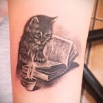 Прелестная татуировка на руке: маленький котенок, увлеченно читающий книгу, рядом с чашкой горячего напитка. Идеально для любителей кошек и чтения.