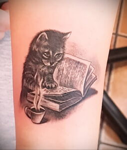 Прелестная татуировка на руке: маленький котенок, увлеченно читающий книгу, рядом с чашкой горячего напитка. Идеально для любителей кошек и чтения.