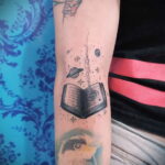 Красивая татуировка на руке: открытая книга с сердцем, окруженная планетами, звездами и космической пылью. Идеально для любителей магии и литературы.