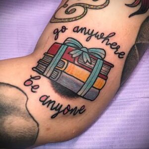 Фото тату на руке: стопка книг, перевязанная голубой лентой, с надписью 'go anywhere be anyone', выполненная в ярком и красочном стиле.