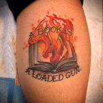 Фото тату на бедре: горящая книга с надписью 'A Book Is A Loaded Gun...', выполненная в стиле олдскул с использованием красного и черного цветов.