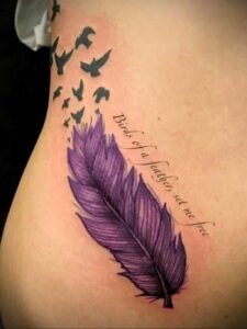 Фото тату на боку: фиолетовое перо с надписью 'Birds of a feather, set me free' и силуэтами птиц, улетающих вверх. Красивый рисунок татуировки для женщин.