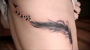 Фото тату на бедре: перо с летящими птицами, выполненное в технике blackwork. Элегантный и минималистичный дизайн.