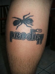 Фото тату: муравей над логотипом группы The Prodigy, выполненный в черном цвете на ноге.