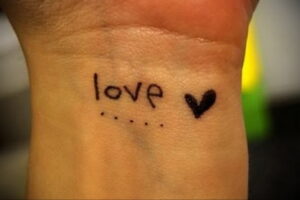Фото тату надписи 'love' с маленьким сердечком на запястье, выполненное черными чернилами в минималистичном стиле.