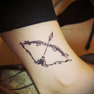 Фото тату на лодыжке: стилизованный лук и стрела, украшенные витиеватыми узорами и листьями. Рисунок выполнен в черных чернилах с высокой детализацией.