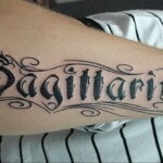 Фото тату на руке с надписью 'Sagittarius' выполненной в каллиграфическом стиле. Элегантный и стильный рисунок татуировки.