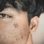 Фото тату маленького разбитого сердца на лице мужчины. Минималистичный и символичный рисунок.