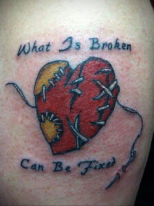 Фото тату разбитого сердца, сшитого черными нитками, с надписью 'What Is Broken Can Be Fixed'. Расположена на плече.