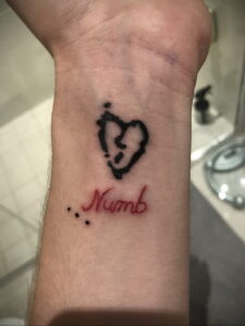 Фото тату разбитого сердца с надписью 'Numb' на запястье. Черный цвет, небрежный стиль, эмоциональный дизайн.