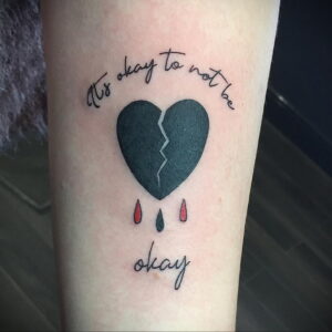 Фото тату разбитого сердца с каплями крови и надписью 'It's okay to not be okay' на предплечье. Черно-красная палитра, реалистичный стиль.