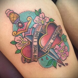 Фото татуировки на бедре: сердце, пронзенное кинжалом, с надписью "For those I love I will sacrifice". Яркие цвета и детализированный дизайн.