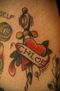Фото татуировки на руке: сердце, пронзенное кинжалом, с именем 'Chloe' на ленте. Рисунок в стиле олд скул.