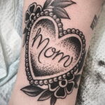 Фото тату на предплечье: сердце, выполненное в технике dotwork, с надписью 'Mom' внутри. Черно-белое изображение, детализированная проработка.