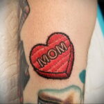 Фото тату на руке: сердце с надписью 'Mom', выполненное в стиле пэчворк. Оригинальный стиль, яркие цвета, текстурная проработка.