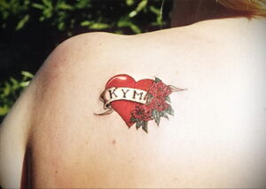 Фото татуировки на плече: красное сердце, обвитое розами, с надписью "KYM" на ленте. Рисунок татуировки в традиционном стиле.