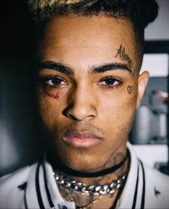 Фотография XXXTentacion с множеством татуировок на лице, шее и голове. Рисунки включают в себя надписи, символы и изображения.