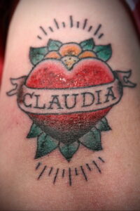Фото тату: традиционная татуировка на руке, изображающая красное сердце с именем 'Claudia' на ленте, окруженное зелеными листьями и лучами света. Старошкольный стиль татуировки.