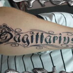 Фото тату надписи 'Sagittarius' на предплечье, выполненной в черном цвете с изящными завитушками и декоративными элементами.