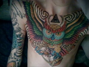 Фото тату совы на груди мужчины, выполненное в ярких и насыщенных цветах с использованием геометрических узоров и символа 'Всевидящее око'.
