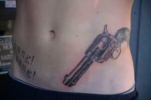 Фото тату револьвера с надписью "Bang Bang" на женском животе. Рисунок выполнен в черно-серых тонах.