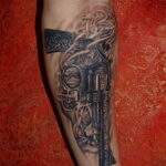 Фото тату: реалистичный рисунок револьвера с клубами дыма и декоративным орнаментом на фоне, выполненный в черно-серых тонах на предплечье.